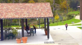 Photographie de la place du village landais, avec un préau équipé de tables et de chaises, dans un environnement verdoyant.